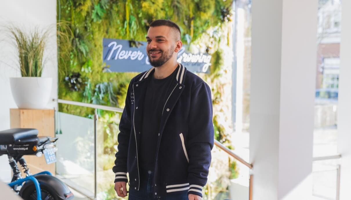 Man in zwart jasje lacht bij planten en bordje 'Stop nooit met groeien', wat zijn passie voor groei bij HOBIJ laat zien.