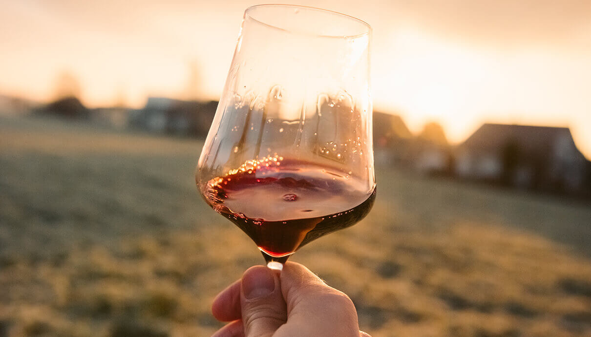 Een hand houdt een Les bulles enchantées-glas met rode wijn vast, dat baadt in de gloed van de zonsondergang boven een wazig landelijk tafereel.