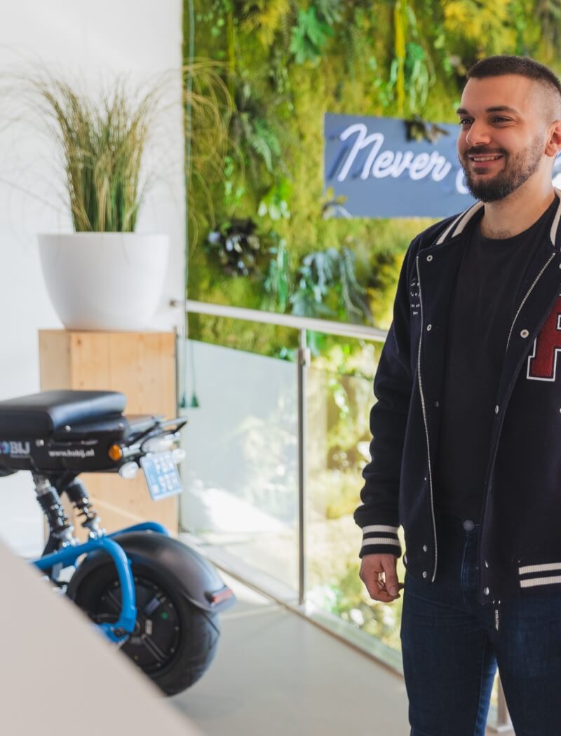 Man lacht binnenshuis bij een blauwe e-bike, groene muur en "Nooit"-bord – TunnelmaDesign-sfeer.