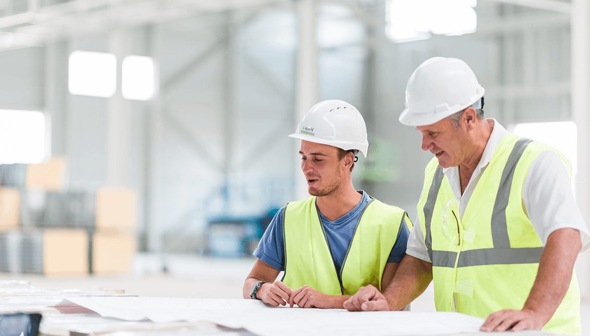 Twee bouwbedrijf Vrolijk-werknemers met veiligheidshelmen en vesten bekijken binnenshuis blauwdrukken.