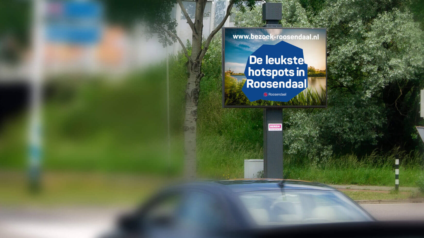 Reclamebord toont "Toplocaties in Roosendaal" met een link naar de website; onscherpe auto op de voorgrond.