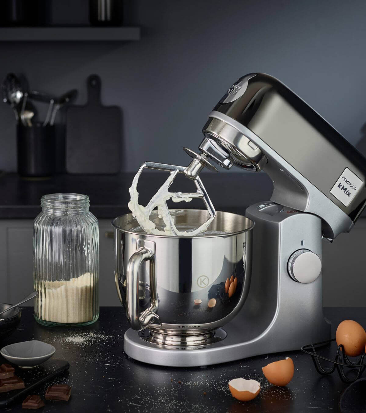 Zilveren mixer met slagroom in een keuken, omringd door eieren, bloem en chocolade.