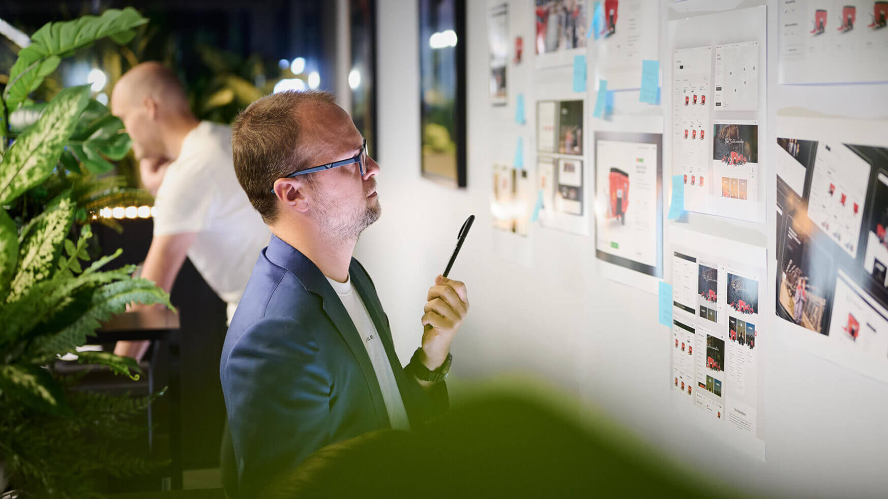 Een man met een bril bekijkt pagina's aan de muur, planten in de buurt. Op de achtergrond bespreekt een collega leadgeneratiestrategieën.