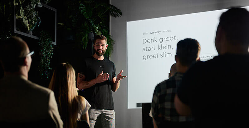 Een presentator legt zijn dia uit aan een schemerige zaal vol luisteraars.