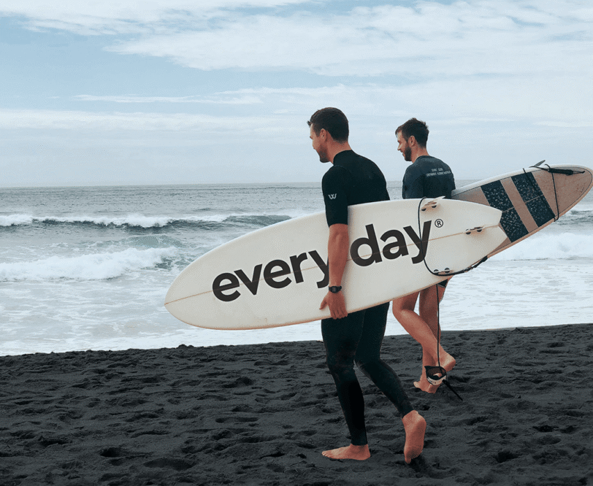 Twee surfers gaan naar de oceaan. De één houdt een bord vast met het opschrift 'elke dag'.