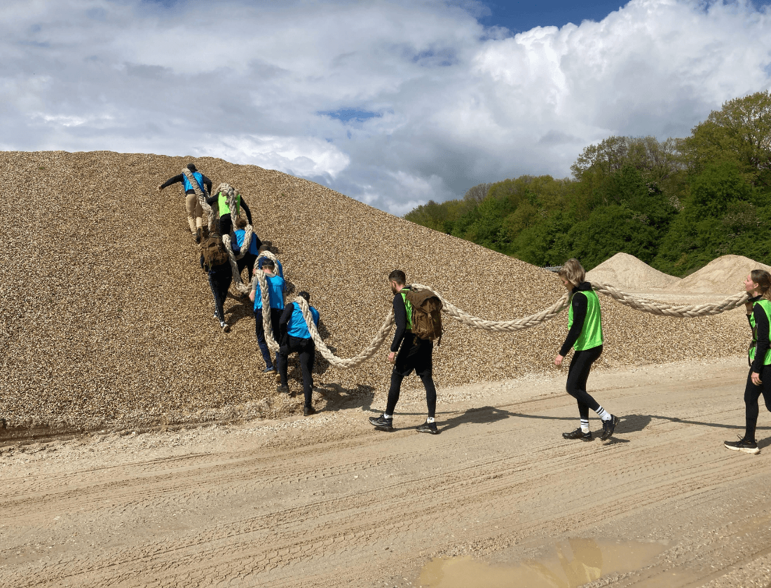 Een groep klimt in één rij een grindheuvel op, allemaal vasthoudend aan hetzelfde dikke touw.