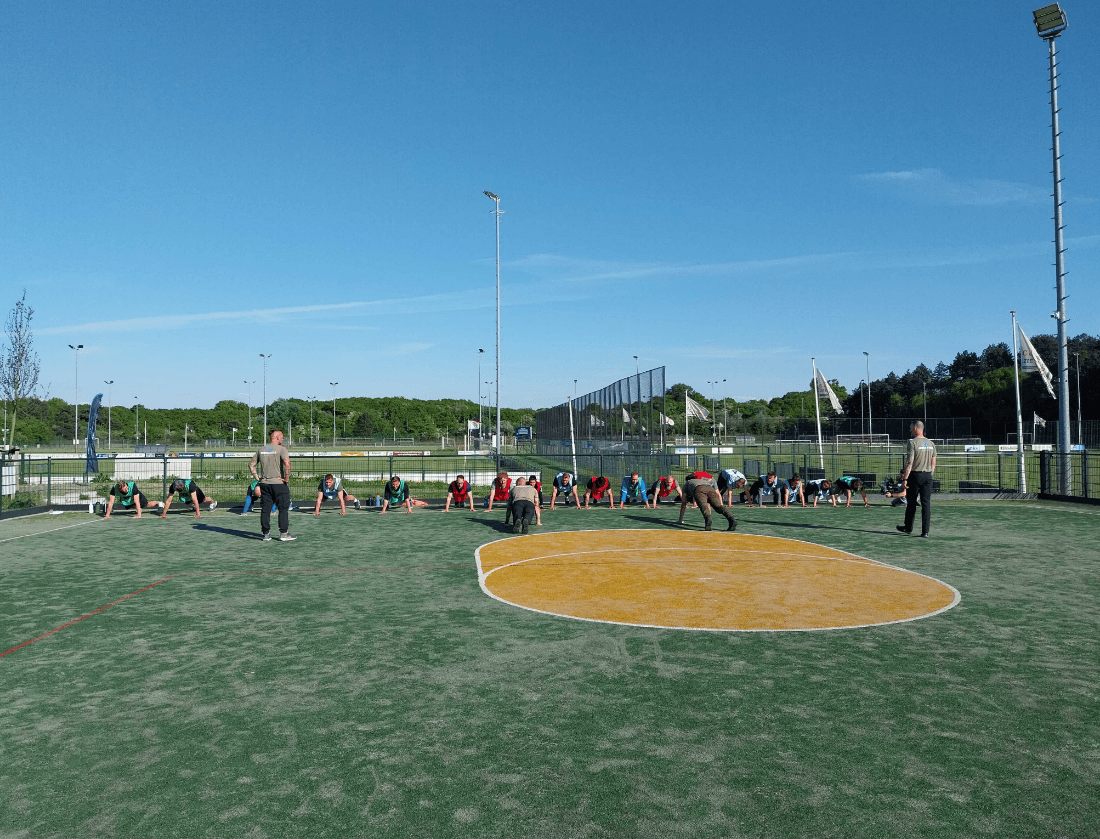 Mensen strekken zich buiten uit op een sportveld met twee instructeurs, onder een strakblauwe lucht.