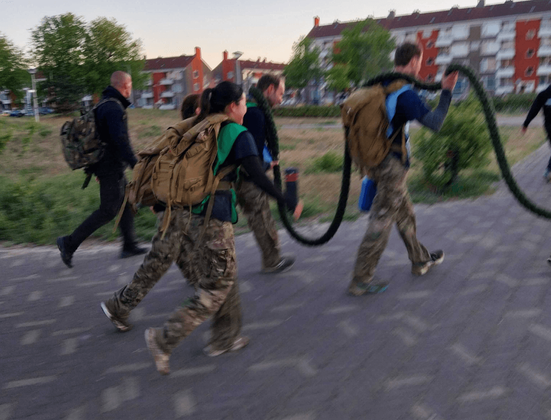 Vijf mensen in camouflagebroeken en rugzakken wandelen samen, terwijl ze een lang, zwaar touw dragen.