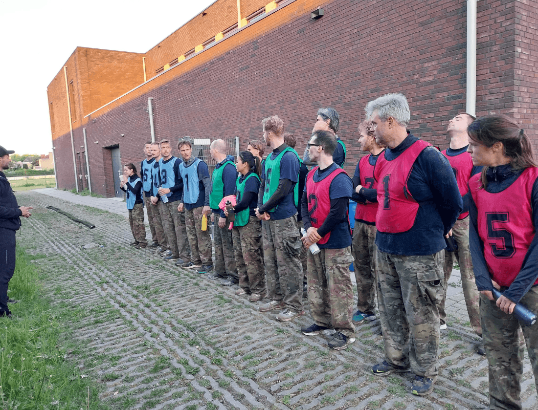 Een groep in camouflagebroeken en vesten luistert naar iemand in het zwart die buiten instructies geeft.