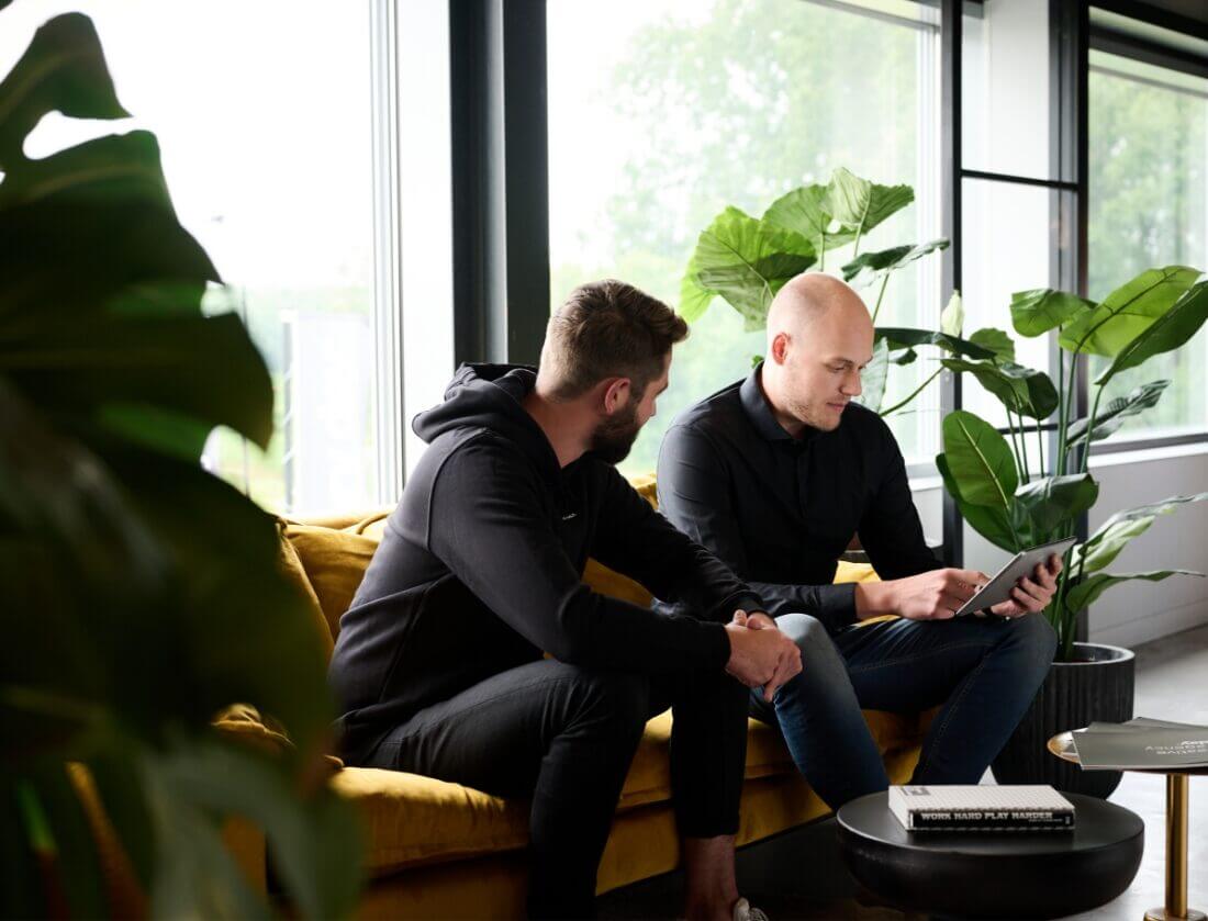 Twee mannen op een gele bank bespreken Magento alternatieven op een tablet in een stijlvol kantoor vol planten.