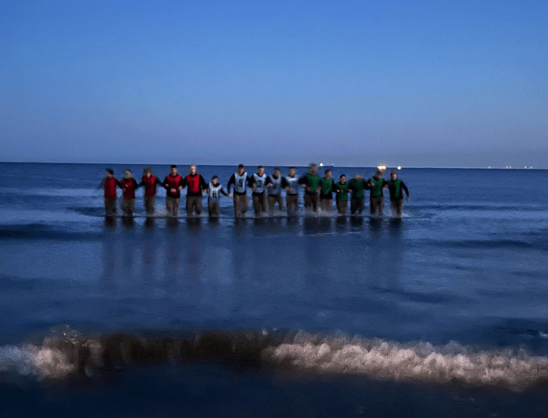 Mensen in felgekleurde shirts staan in ondiep zeewater bij zonsondergang, onder een blauwe lucht.