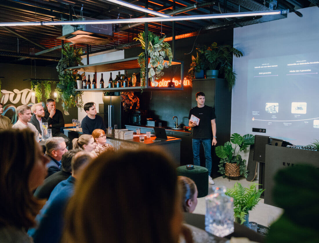 Een man presenteert aan een zittende groep in een stijlvolle, met planten gevulde ruimte met een groot scherm.