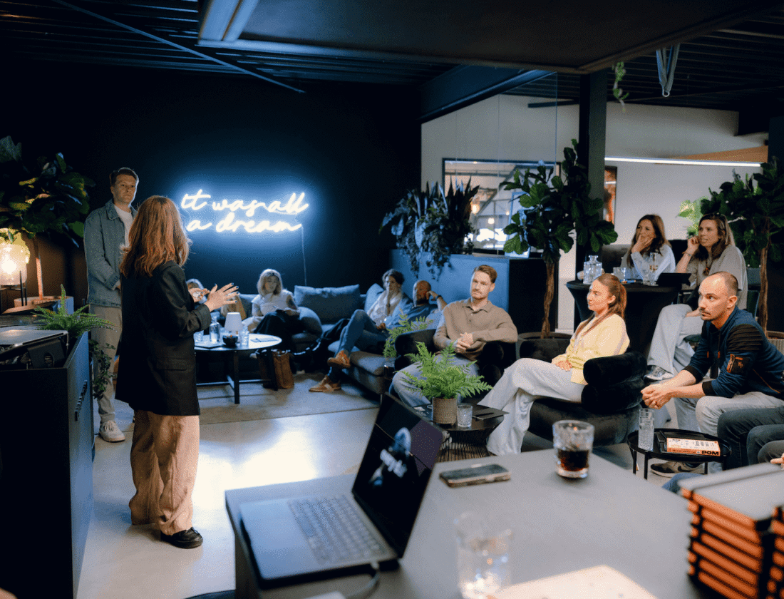 Een vrouw presenteert aan een zittende groep in een modern kantoor met een neonbord: "het was allemaal een droom.
