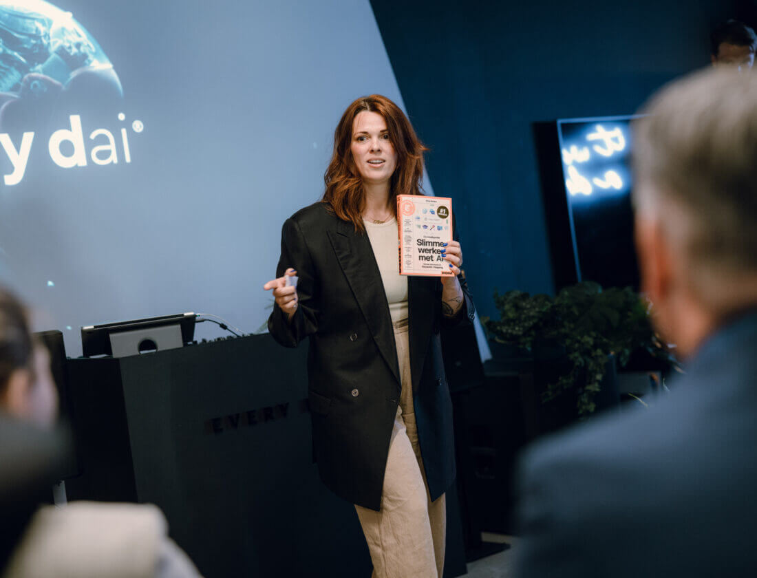 Vrouw presenteert aan een publiek, houdt een boek vast, met een presentatiescherm op de achtergrond.
