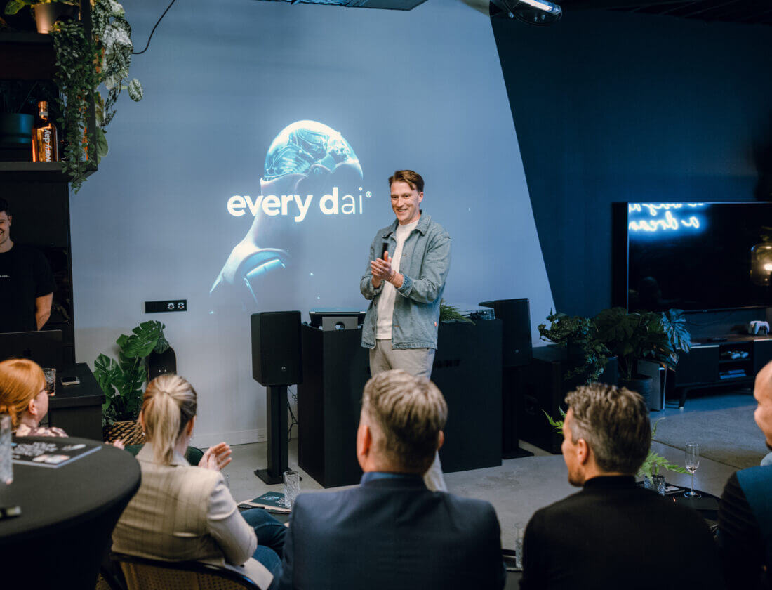 Man spreekt een publiek toe in een strakke kamer, met "every dai" achter hem geprojecteerd.