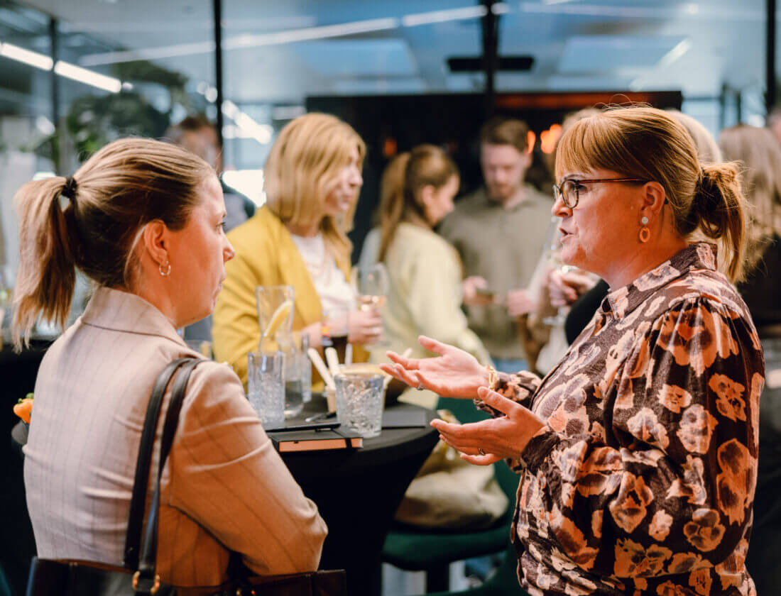 Twee vrouwen chatten op een levendig netwerkevenement, met pratende groepen en drankjes op tafels op de achtergrond.