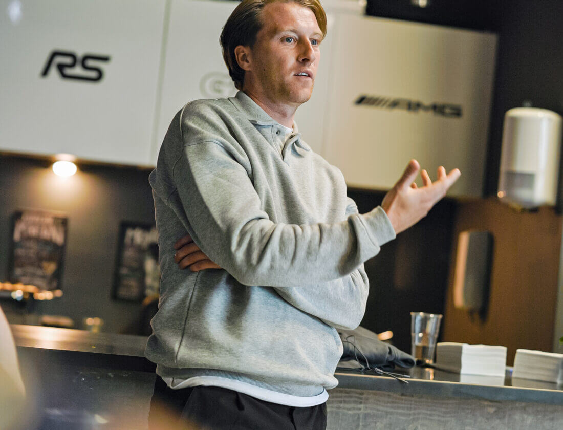 Man met grijze capuchon gebaart terwijl hij spreekt in een strakke, moderne keuken of café.