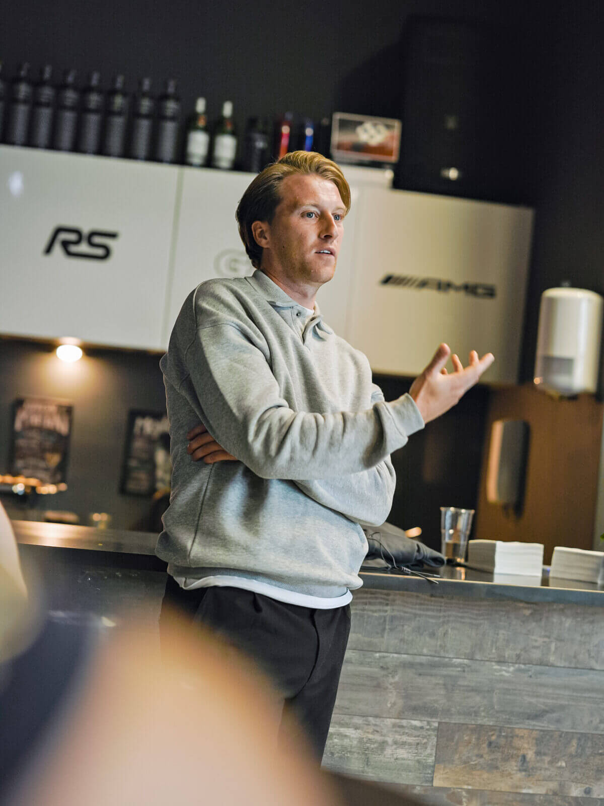 Man met grijze capuchon gebaart terwijl hij spreekt in een strakke, moderne keuken of café.