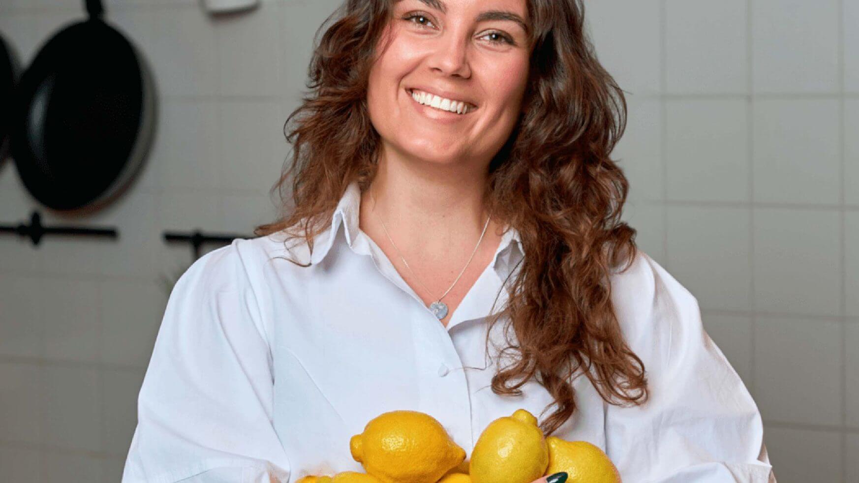 Vrouw in een wit shirt glimlacht, houdt citroenen vast in een keuken met betegelde muren en hangende pannen.