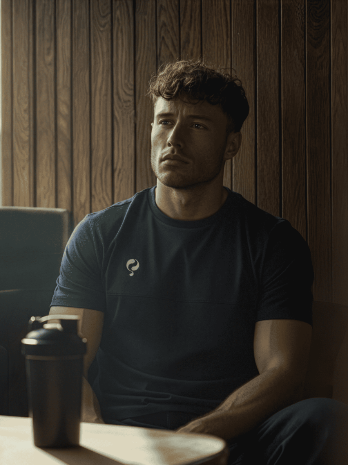 Man in donker T-shirt zit binnenshuis bij een houten wand, shakerfles op de tafel voor hem.