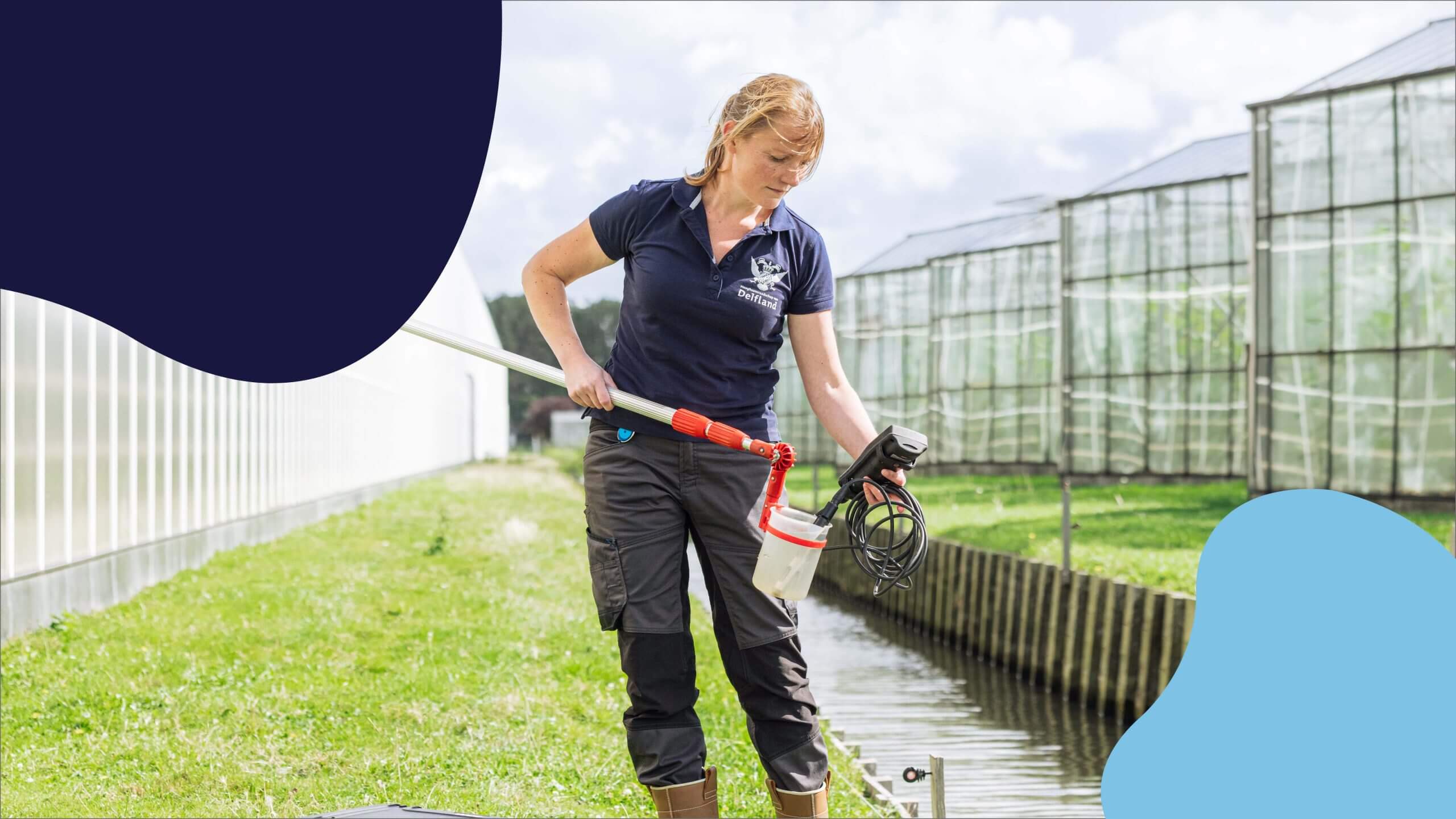 Vrouw in werkkleding bemonstert water uit een kanaal naast kassen op een zonnige dag.