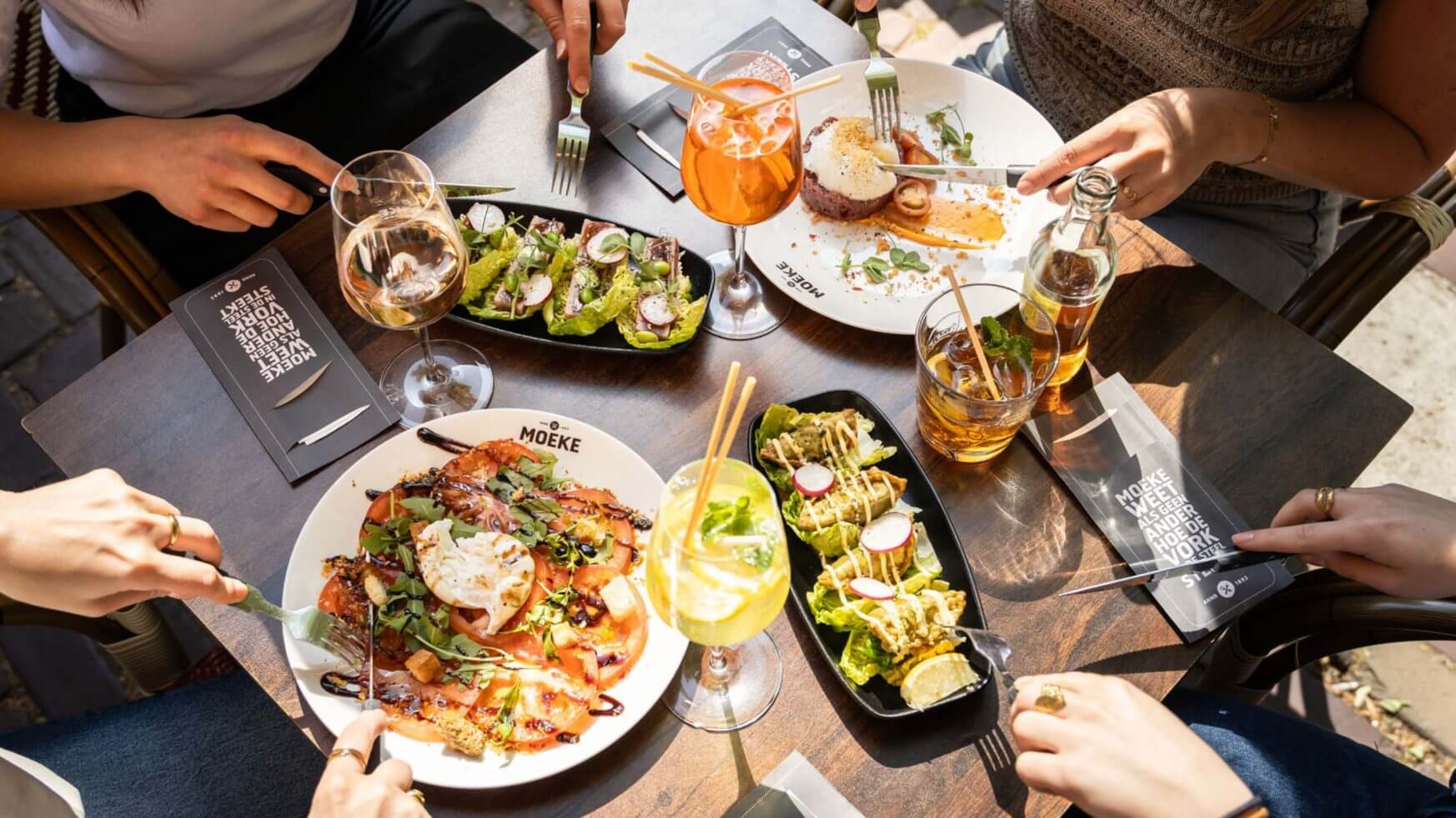 Vier vrienden delen salades, drankjes en hapjes aan een zonnige tafel in een buitenrestaurant.