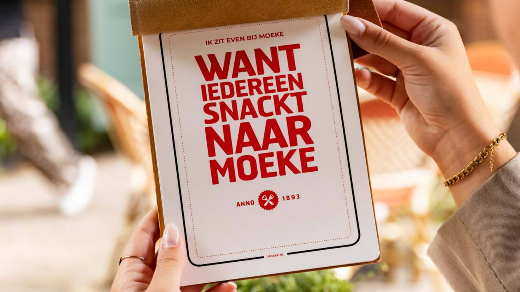 Persoon die een menukaart vasthoudt met vette rode Nederlandse tekst: "WANT IEDEREEN SNACKT NAAR MOEKE.