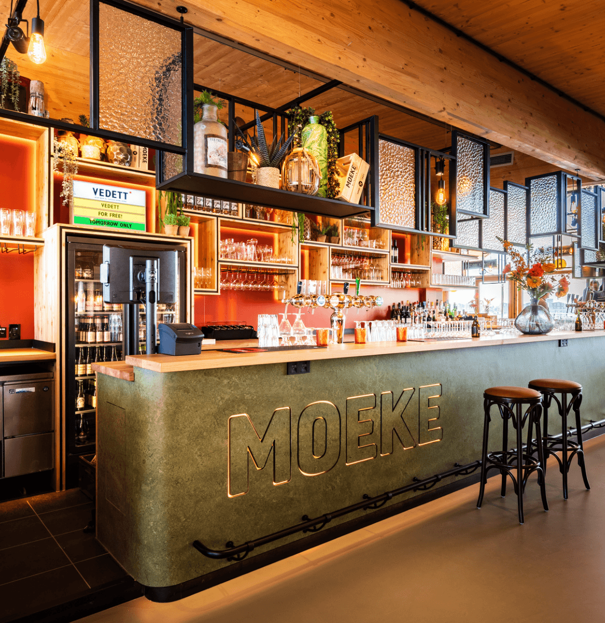 Chique bar met een groene MOEKE toonbank, planken met flessen en gezellige sfeerverlichting.