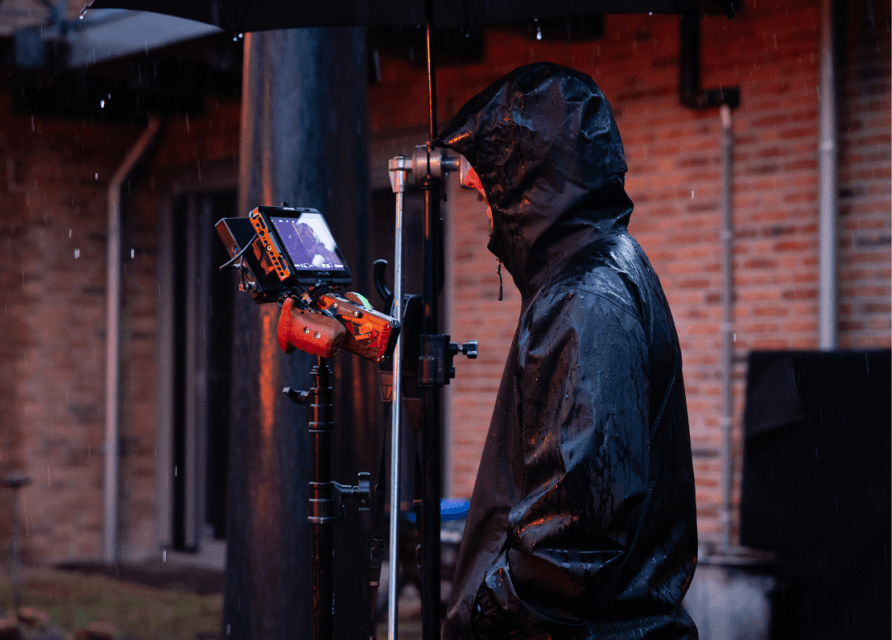 Persoon in regenjas onder paraplu met een cameramonitor buiten op een regenachtige dag.
