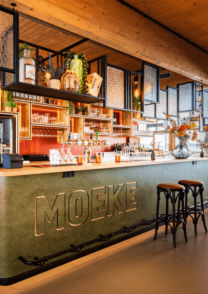 Chique bar met MOEKE-bord, rekken met flessen en glazen en een eigentijdse inrichting met groen.
