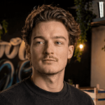 Jonge man met golvend bruin haar en een snor kijkt opzij in schemerig binnenlicht.