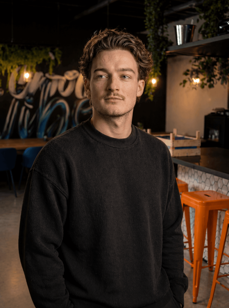 Man in een donkere trui in een stijlvol café met planten en oranje krukjes op de achtergrond.