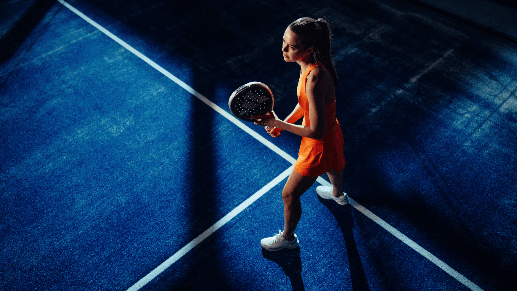 Een vrouw in een oranje jurk met een padelracket staat klaar op een blauwe XNRGY-baan.
