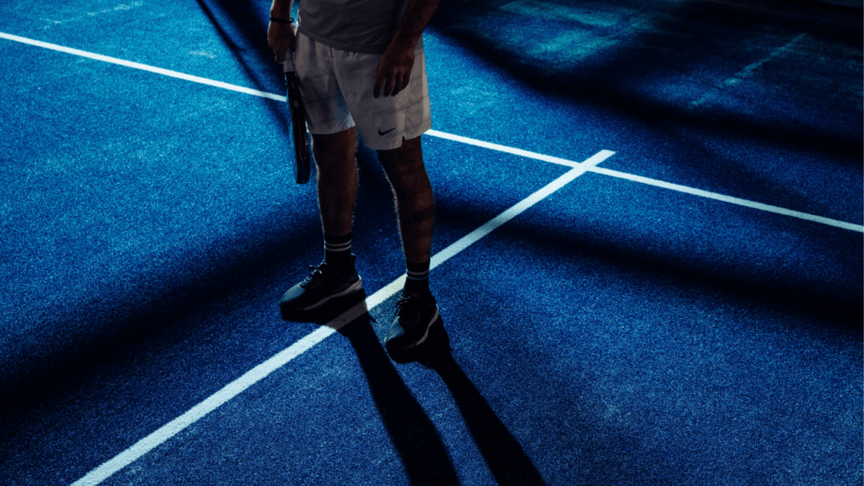 Een persoon op een blauwe tennisbaan met een racket in de hand, met uitgesproken schaduwen en dramatische XNRGY-verlichting.