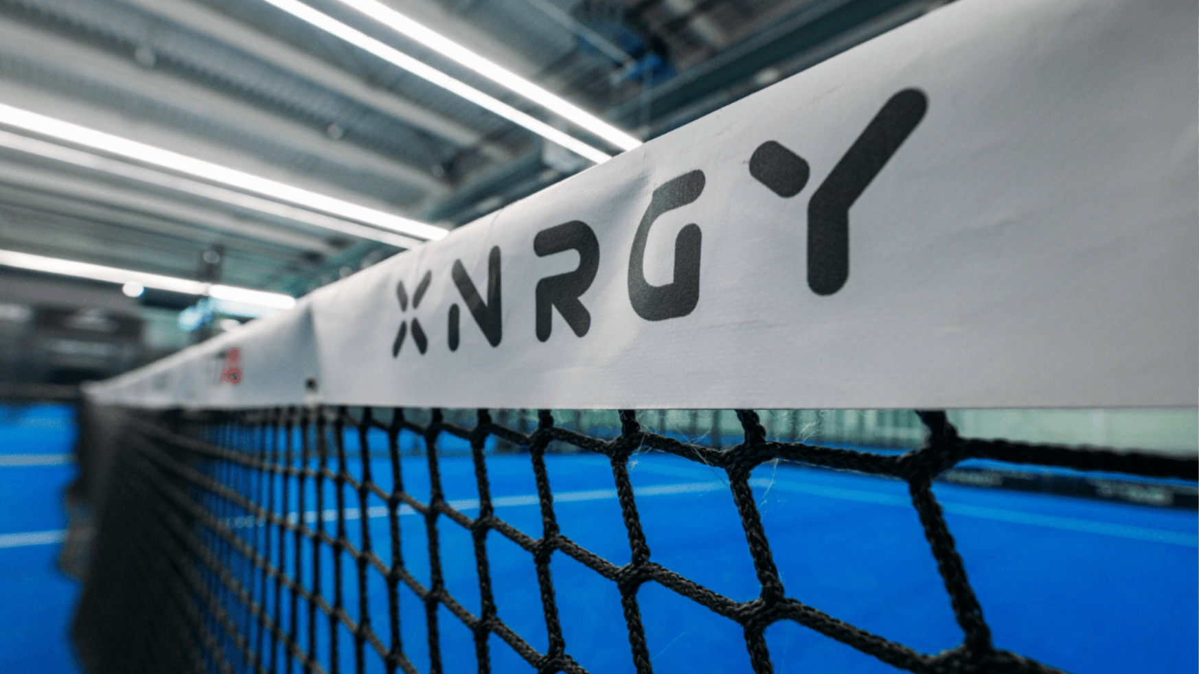 Close-up van een tennisnet met XNRGY op witte tape, met op de achtergrond een blauwe indoorbaan.