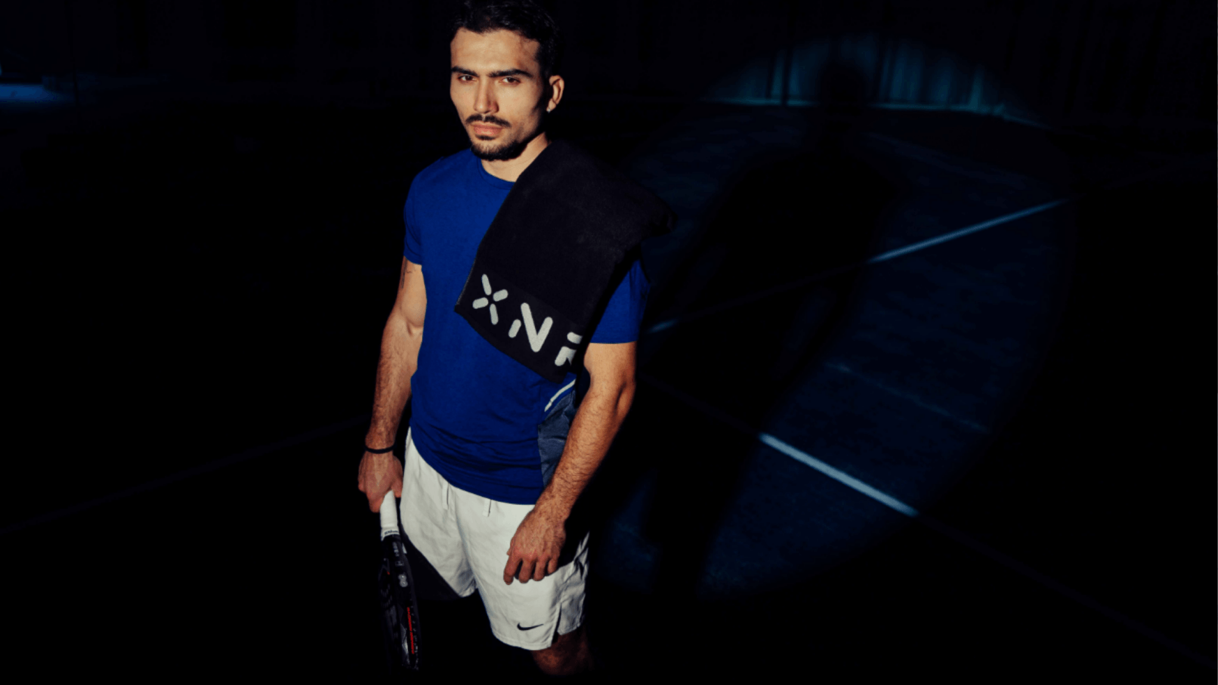 Een man in een blauw XNRGY-shirt en een witte korte broek staat met een racket in zijn hand op een donkere tennisbaan, met een handdoek over zijn schouder geslagen.