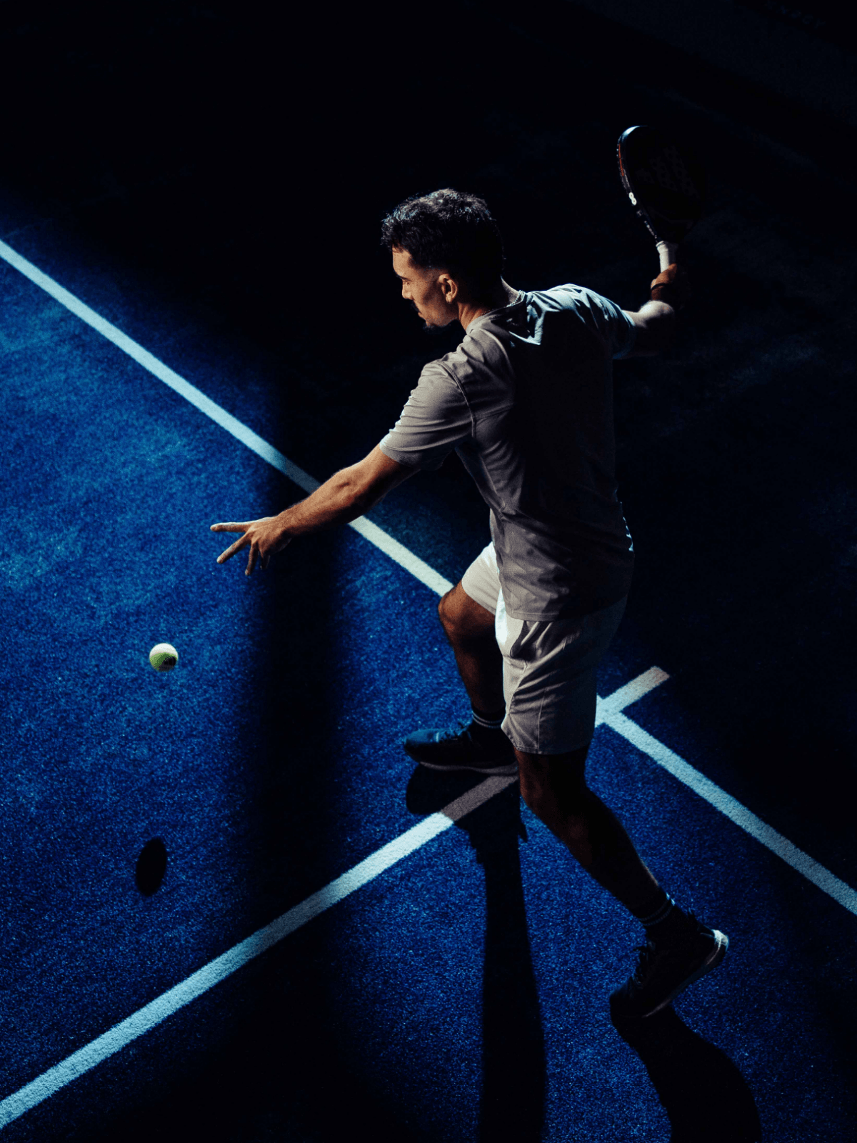 Een tennisser staat klaar om uit te halen op een blauwe XNRGY-baan onder opvallende, dramatische verlichting.