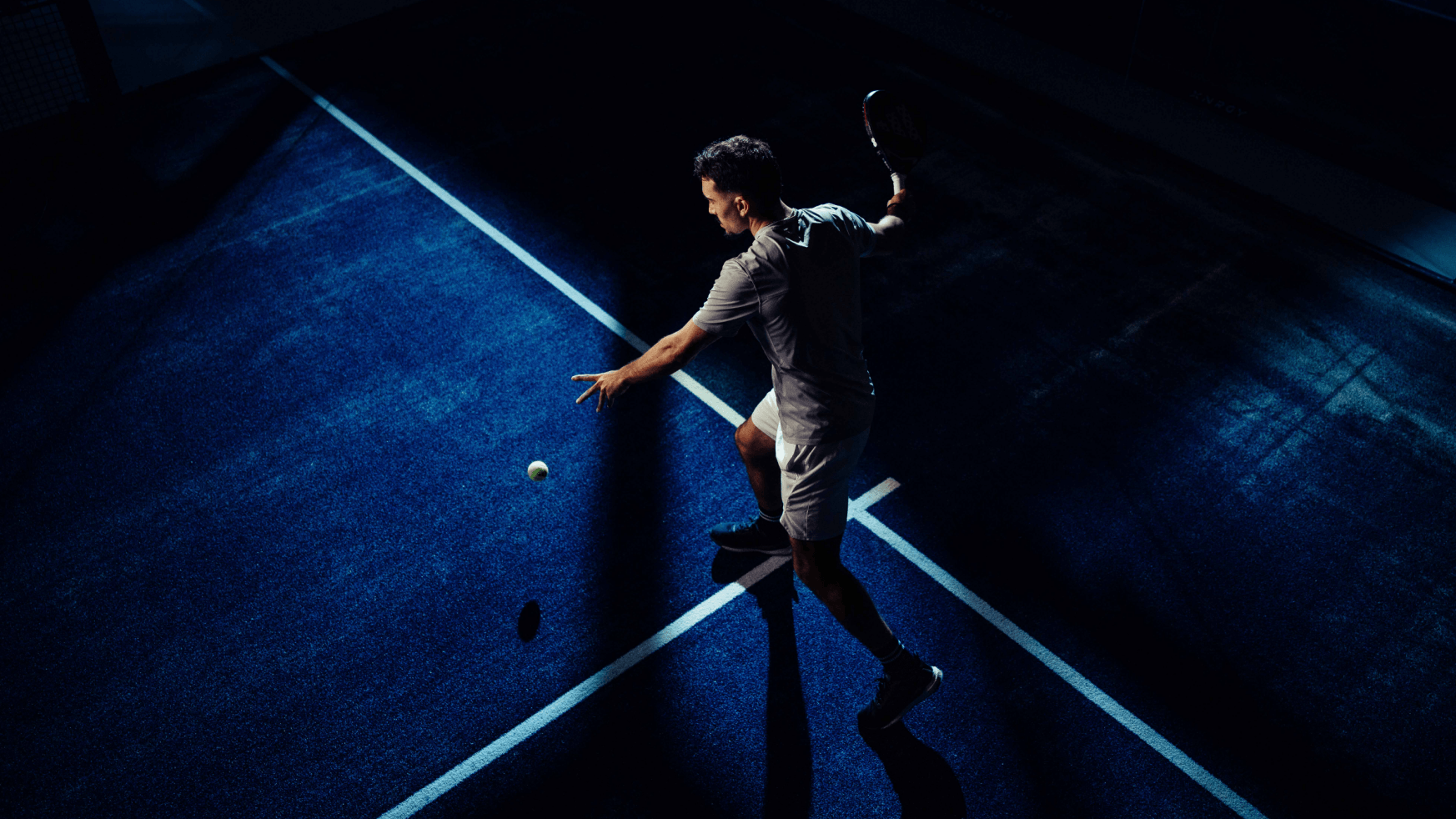 Een tennisser staat klaar om uit te halen op een blauwe XNRGY-baan onder opvallende, dramatische verlichting.