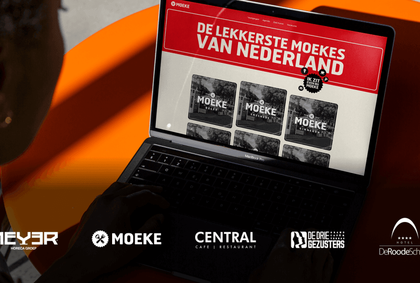 Een laptopgebruiker die aan een oranje tafel de website ‘De Lekkerste Moekes van Nederland’ bekijkt.