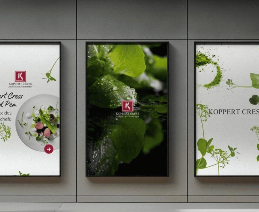 Drie posters met groene bladeren en het Koppert Cress-logo aan de muur; twee wazige figuren lopen voorbij.