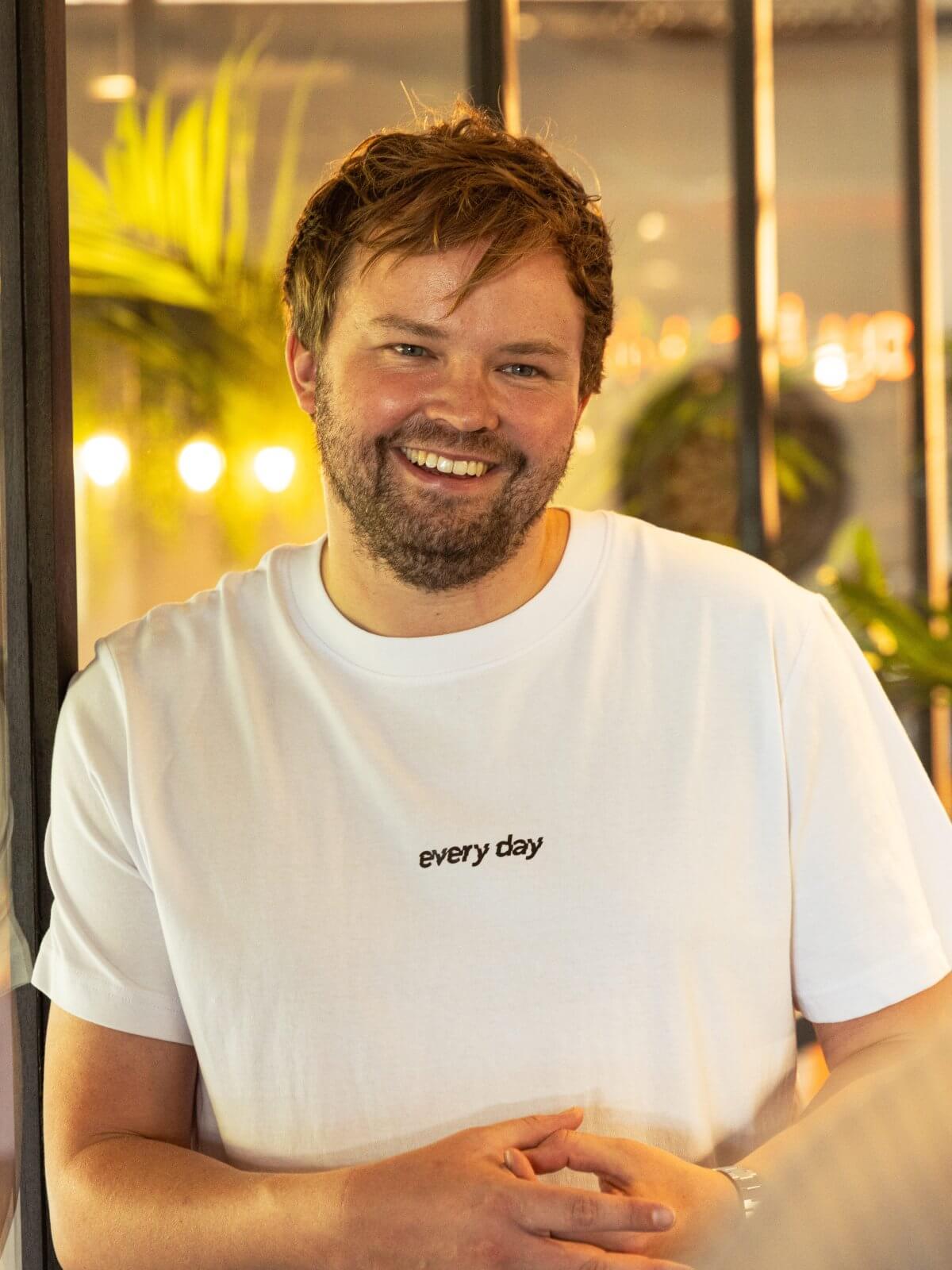 Een man in een wit T-shirt die glimlacht terwijl hij met een collega praat op kantoor.