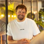 Een man in een wit T-shirt die glimlacht terwijl hij met een collega praat op kantoor.