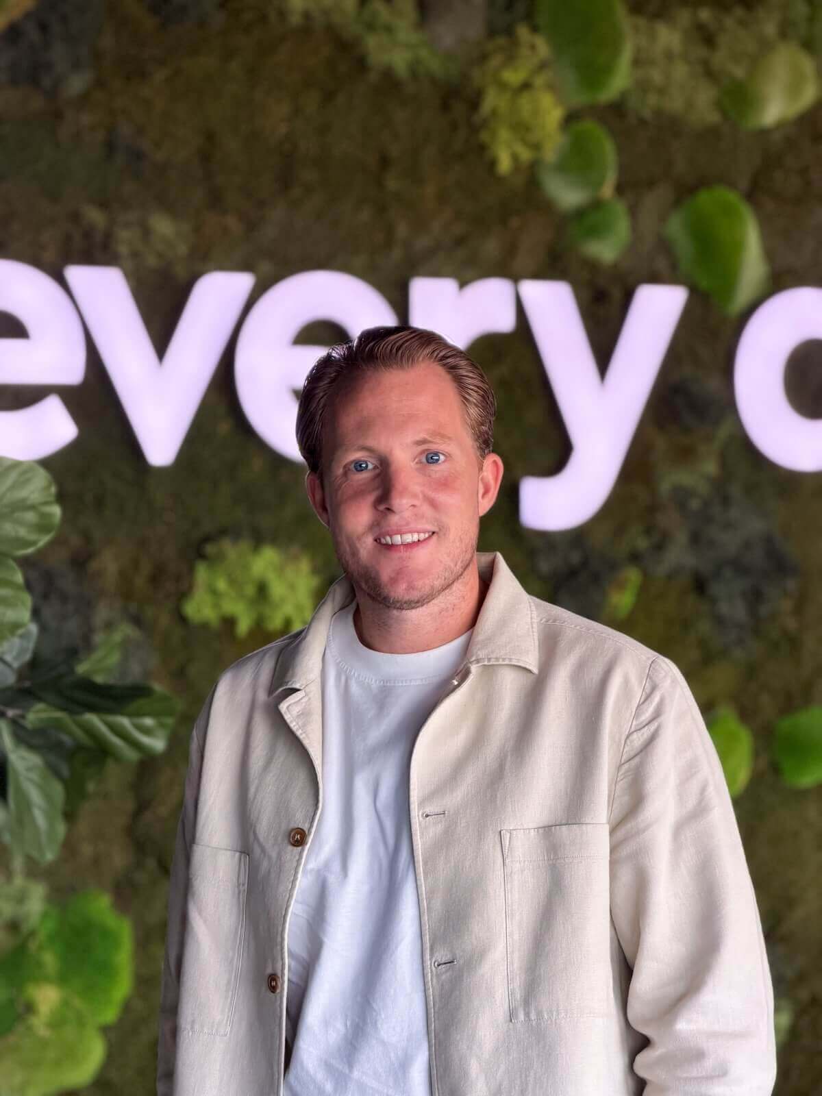 Een glimlachende man in een licht jasje die poseert voor een groene plantenwand met verlichte tekst.