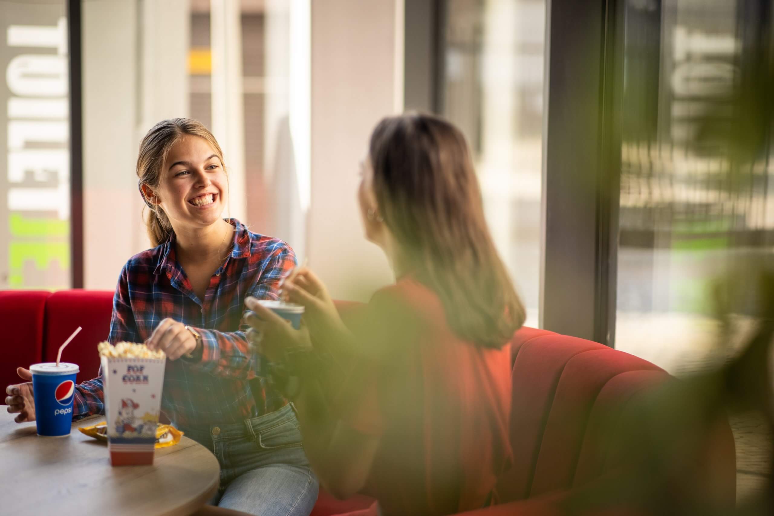 Twee vrouwen lachen en kletsen gezellig aan een zonnig cafétafeltje, onder het genot van popcorn en een drankje.