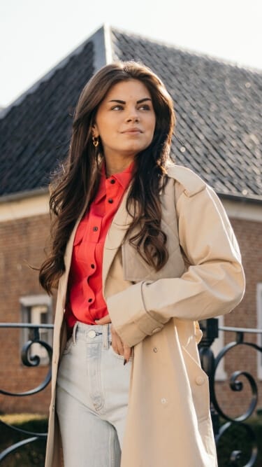 Een vrouw in een beige jas en een rood shirt staat bij een hek, met achter haar een bakstenen gebouw.