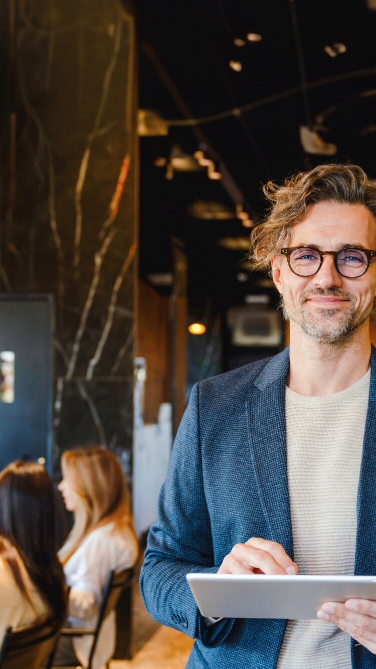 Een lachende man met een bril houdt een tablet vast in een druk, modern café.