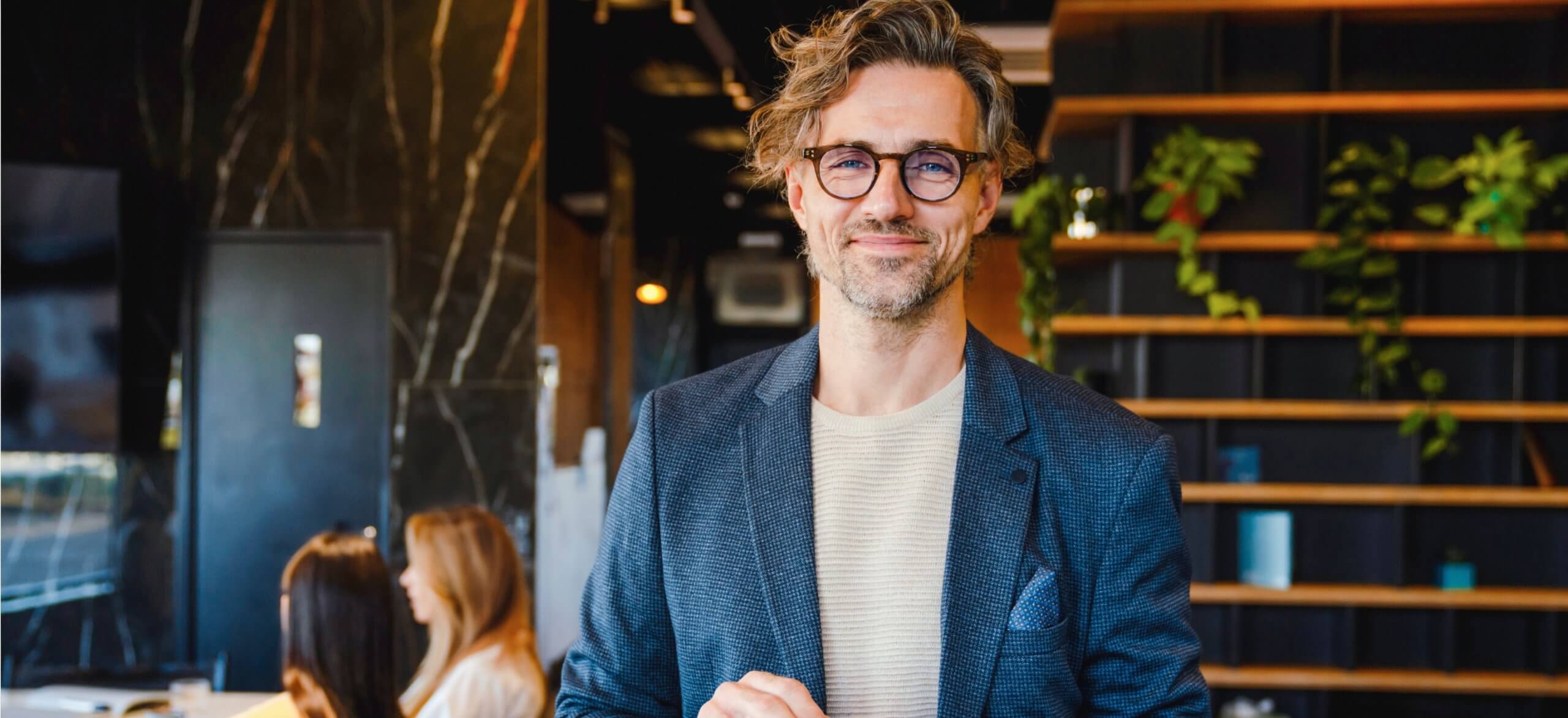 Man met bril lacht in modern kantoor, blazer aan, omringd door planten en collega's.