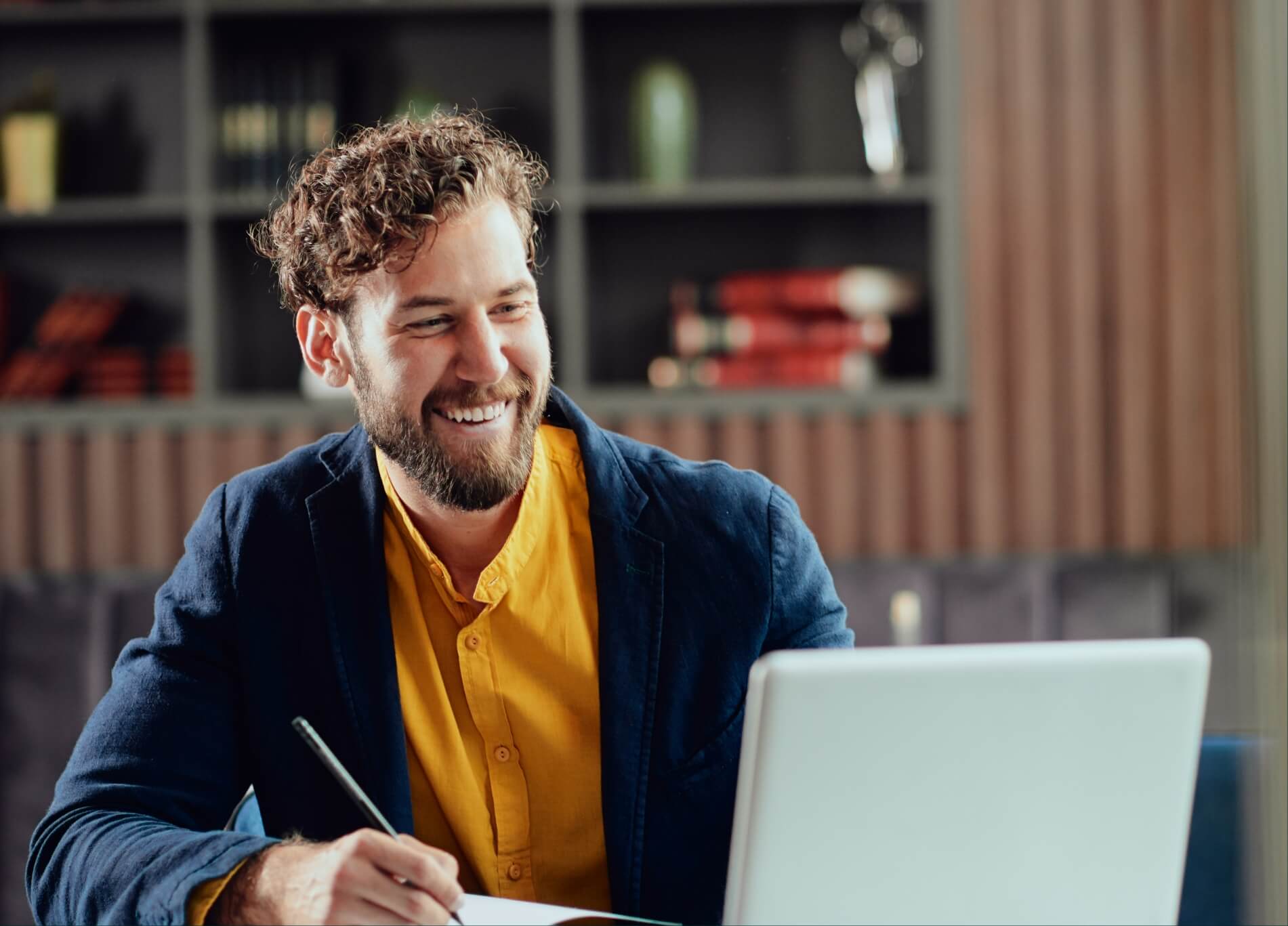Lachende man in een geel overhemd en een blauw jasje typt op zijn laptop en schrijft met een pen in een modern kantoor.