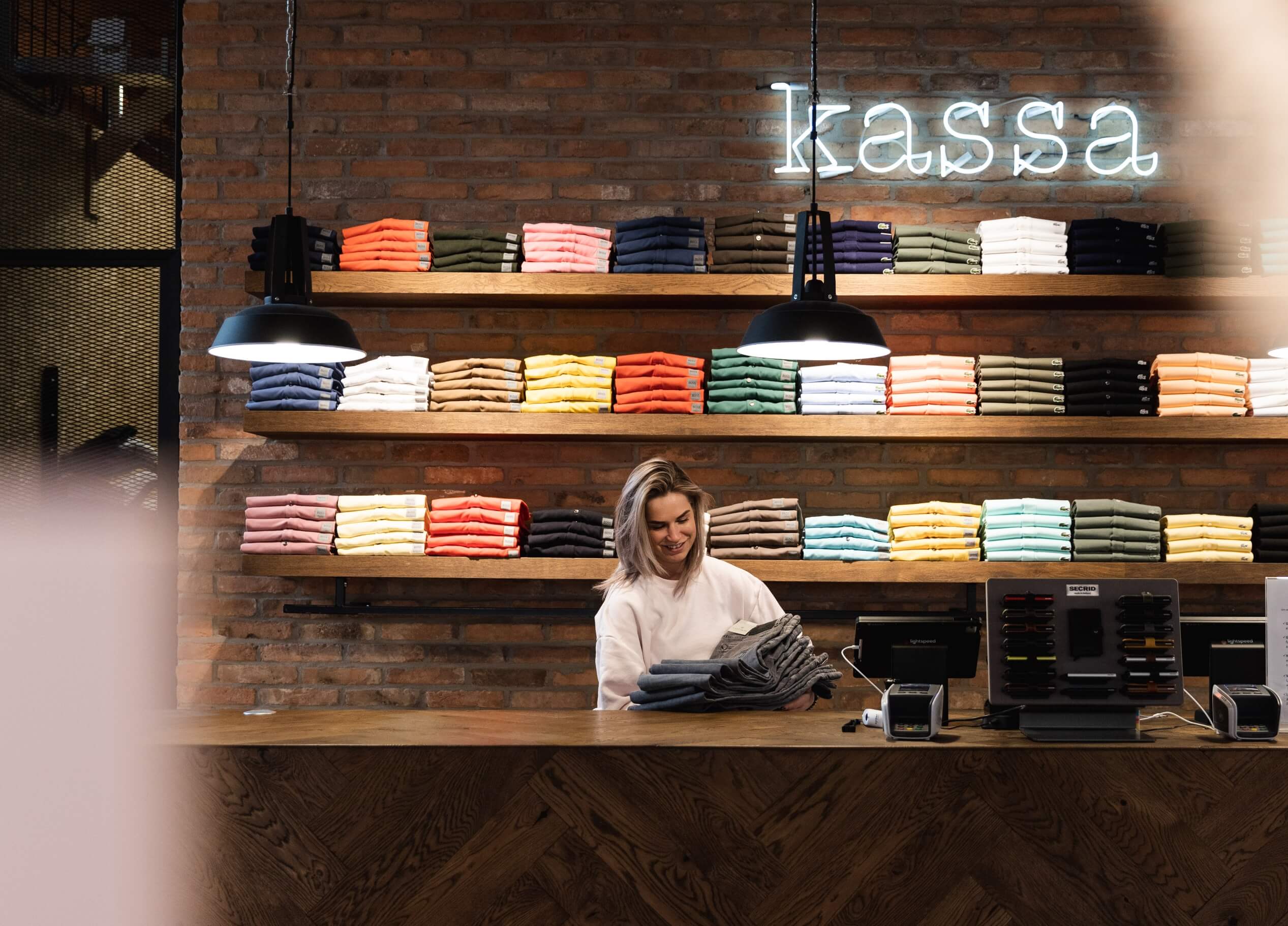 De kassier vouwt kleding op achter een houten toonbank met daarop kleurrijke overhemden, onder een bordje met de tekst 'Kassa'.