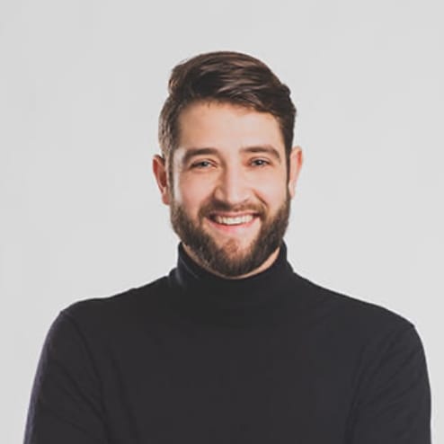 Een man met een baard en een donkere coltrui glimlacht tegen een lichte achtergrond.